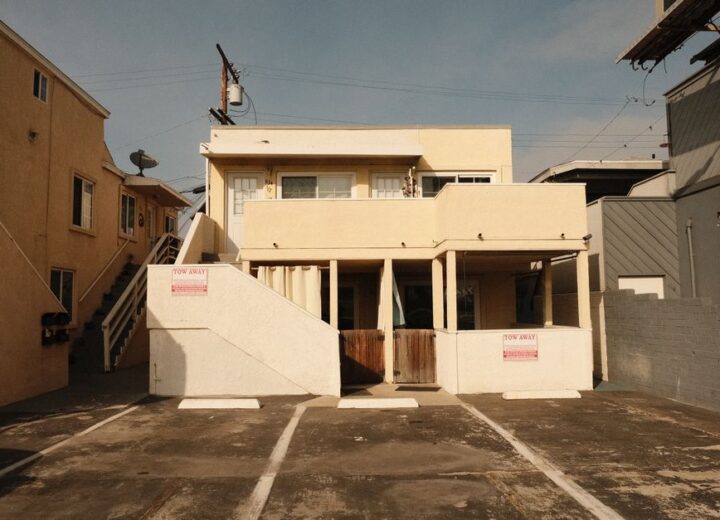 A two-story beige concrete building with a flat roof showing a residential architecture style, captured in an empty driveway.