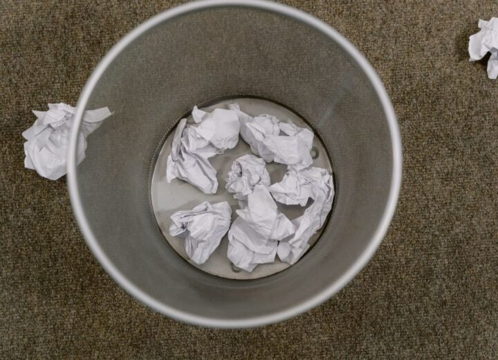 Overhead view of a trash can filled with crumpled paper on a carpeted floor.