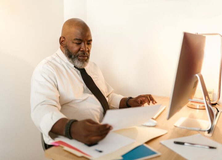 Bald man with beard in white shirt reviewing documents at desk with computer.