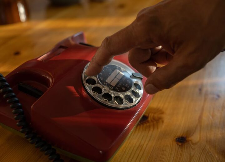 Close-up of a hand dialing a red rotary phone on a wooden table.
