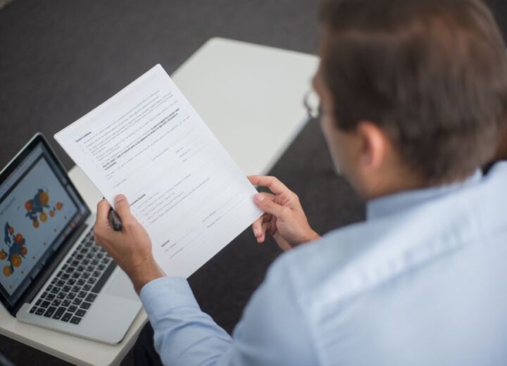 Professional in blue shirt reviewing documents next to laptop displaying charts.