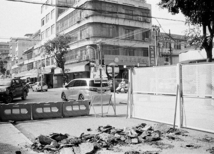 Street view of an urban construction site with barrier fencing and passing vehicles.